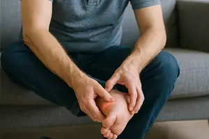 Middle-aged man sitting on a sofa, grimacing in pain while examining the sole of his bare foot, symbolizing diabetic neuropathy discomfort.