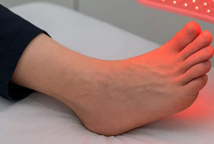 A medical professional in a white lab coat applies red and infrared light therapy to a patient’s foot using a handheld LED device during a clinical treatment session for peripheral neuropathy